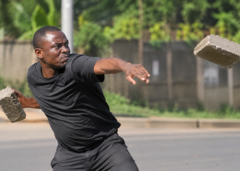 Illustrative photo: A man throws a concrete paver in a simulated attack. The image is used for illustration and does not depict the actual incident involving Minister Justine Nameere’s driver.