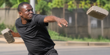 Illustrative photo: A man throws a concrete paver in a simulated attack. The image is used for illustration and does not depict the actual incident involving Minister Justine Nameere’s driver.